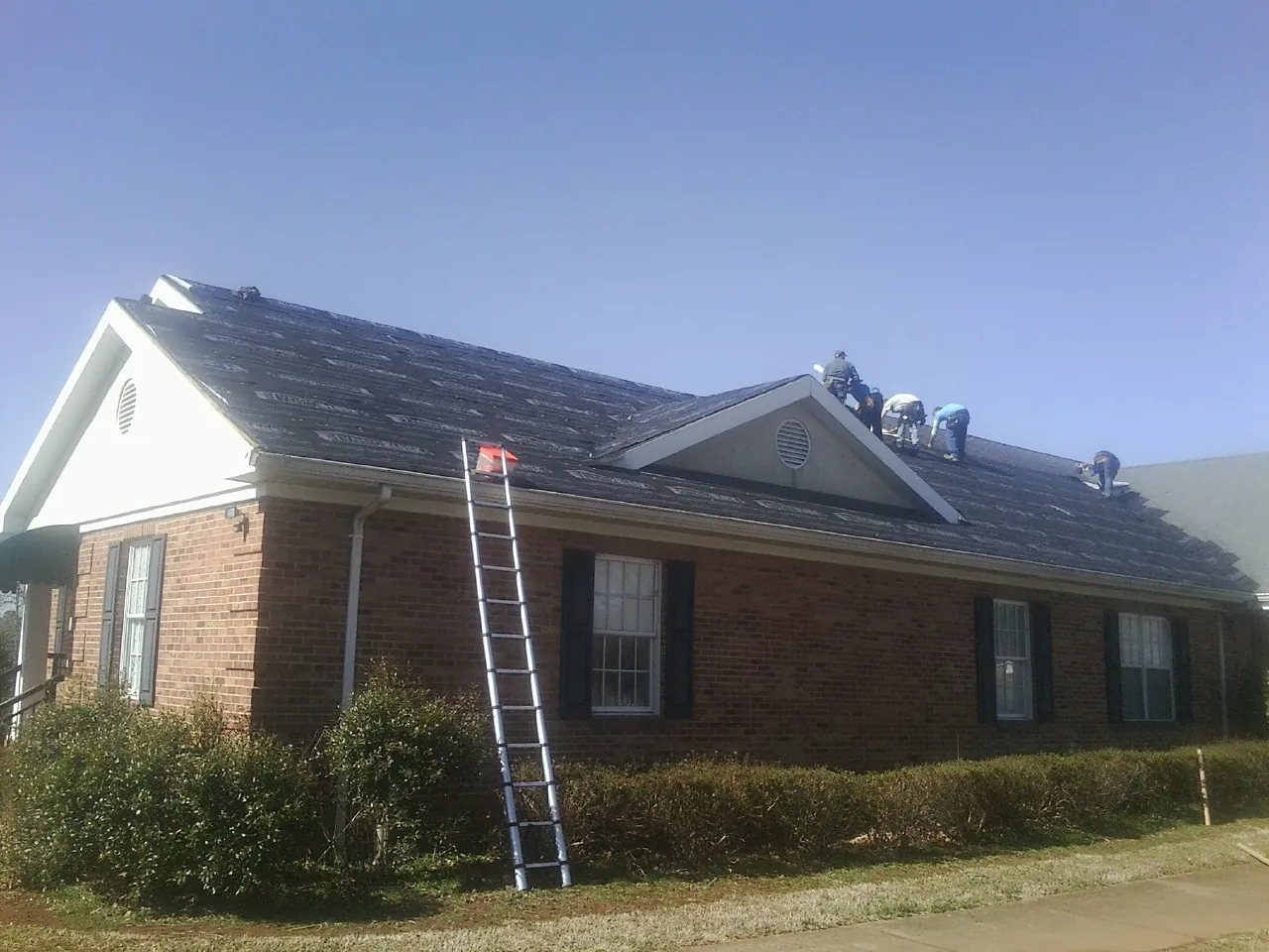Crew tearing shingles off a brick ranch roof with a ladder set — Metal Roof Repair work by our crew in Soldier