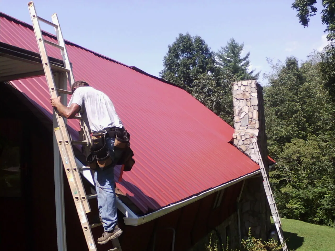 Roofer on a ladder beside a red metal roof and stone chimney — Soffit & Fascia Repair by Roofing Brothers in Soldier